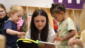 A high school student visiting Corse for a Family and Education CTE Pathway class reads a book to two toddlers in Corse Early Childhood Center's childcare program.