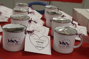 Two rows of mugs displaying the logo for Club M youth mentoring program and a note that says "Everything is s'more fun at Club M" sit on a table