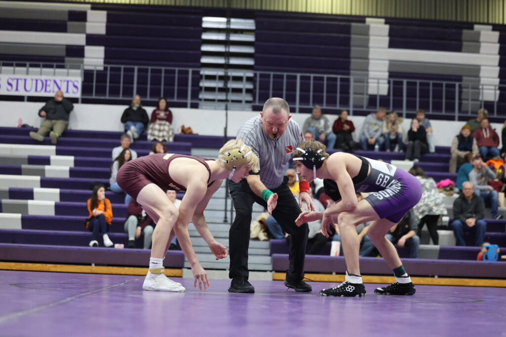 A Burlington wrestler and his opponent crouch face-to-face on the wrestling mat as the referee signals for their round to begin.