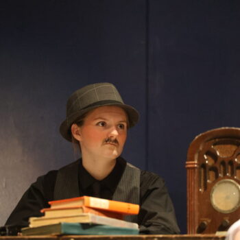 A student with a drawn on mustache and bowler hat looks worried while sitting at a table atop which sit a pile of books and a clock during dress rehearsal for the spring play, 