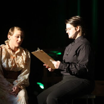 A student actor holds a clipboard while her fellow cast member speaks and a green light glows in the background during the spring play, 