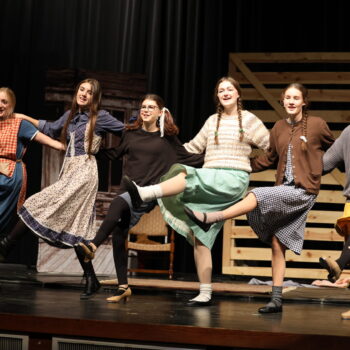 Seven student actresses stand in a line while doing a leg kick during the fall production of 