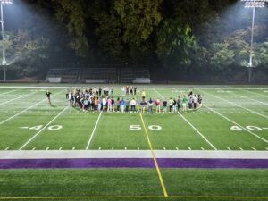 Fellowship of Christian Athletes members stand in a semi-circle in the middle of the football field at Bracewell Stadium.