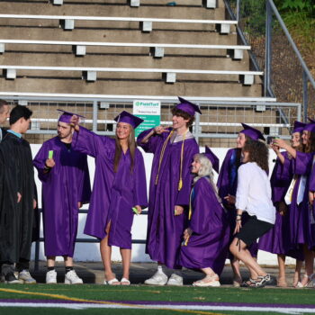 A group of students wearing purple caps and gowns pose for a photo alongside their teacher, Annette Mosbach, before the start of the 2024 commencement ceremony at Bracewell Stadium.