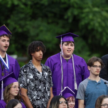 Graduating seniors dressed in purple caps and gowns sing alongside younger choir members during their graduation ceremony at Bracewell Stadium.