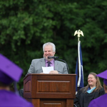 School board president Darven Kendell speaks into a microphone while standing behind a podium during the 2024 Commencement Ceremony at Bracewell Stadium.