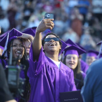 A young man wearing sunglasses holds up his phone to get video and/or photo of himself and the young woman next to them as they turn their tassels during the 2024 commencement ceremony at Bracewell Stadium.