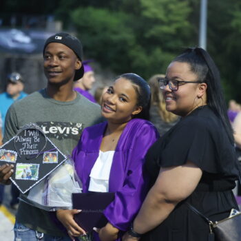 A girl smiles while standing between her parents as her father holds her graduation cap reading, 