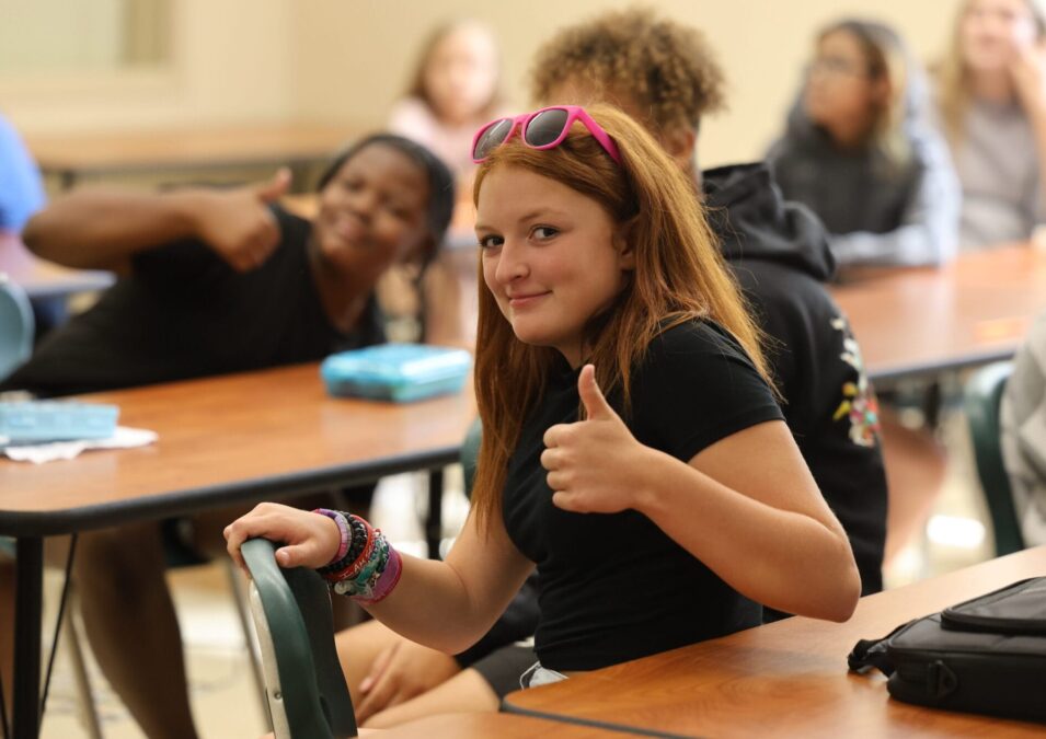 A middle school student gives a thumbs up while sitting at a table in her classroom on the first day of school.