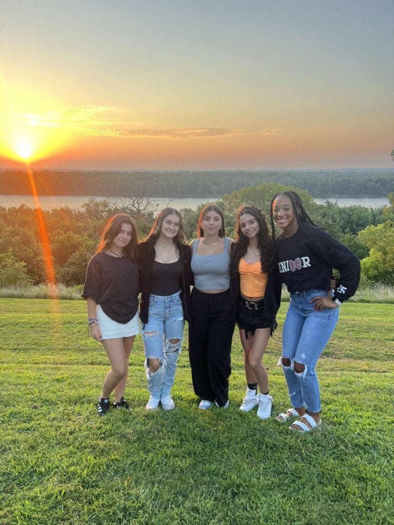 Natalia Lopez Gonzalez and four of her friends smile while posing for a photo outside with trees, the river and sunset in the background.