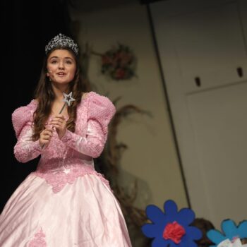 A high school student playing the role of Glinda the Good Witch sings on stage amid a backdrop of flowers during the musical production of 