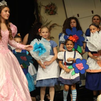 A high school student playing the role of Glinda the Good Witch sings on stage alongside elementary students dressed as munchkins amid a backdrop of flowers during the musical production of 