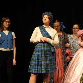 The mayor of Munchkin City sings onstage alongside Glinda the Good Witch and other munchkins during a high school musical production of 