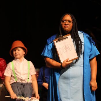 A munchkin coroner holds up a clipboard containing a piece of paper that reads 