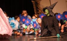 Munchkins peer out from behind stage prop flowers as the Wicked Witch of the West mourns the death of her sister during the high school musical production of 