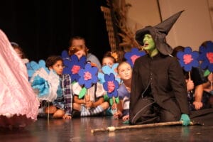 Munchkins peer out from behind stage prop flowers as the Wicked Witch of the West mourns the death of her sister during the high school musical production of "The Wizard of Oz."