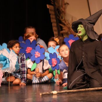 Munchkins peer out from behind stage prop flowers as the Wicked Witch of the West mourns the death of her sister during the high school musical production of 