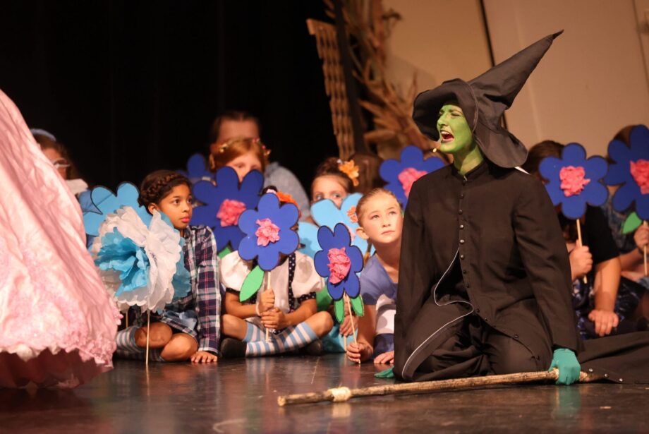 Munchkins peer out from behind stage prop flowers as the Wicked Witch of the West mourns the death of her sister during the high school musical production of 