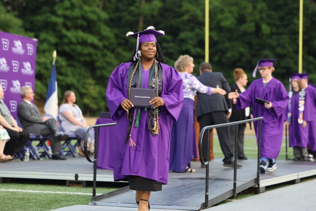 A young woman wearing a purple cap and gown along with many cords representing various accomplishments smiles while holding her diploma after exiting the stage at Bracewell Stadium.