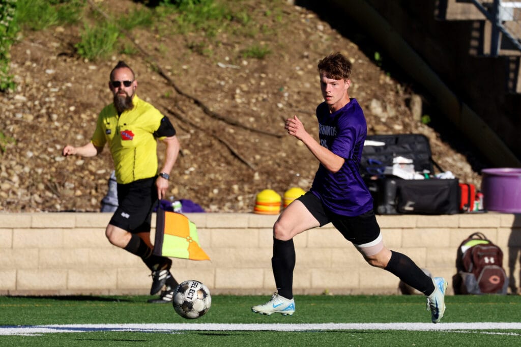 A member of the boys soccer team runs after a ball as a referee follows along in the background.