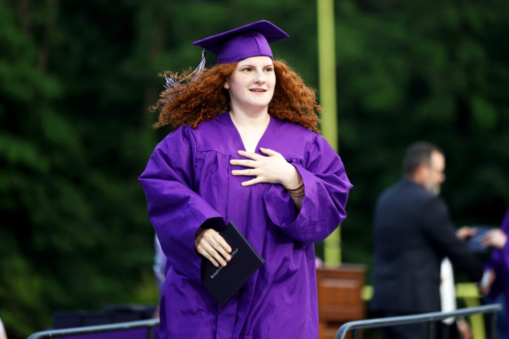 A graduate wearing a purple cap and gown smiles while holding her diploma and returning to her seat during Burlington High School's 2025 commencement ceremony at Bracewell Stadium.