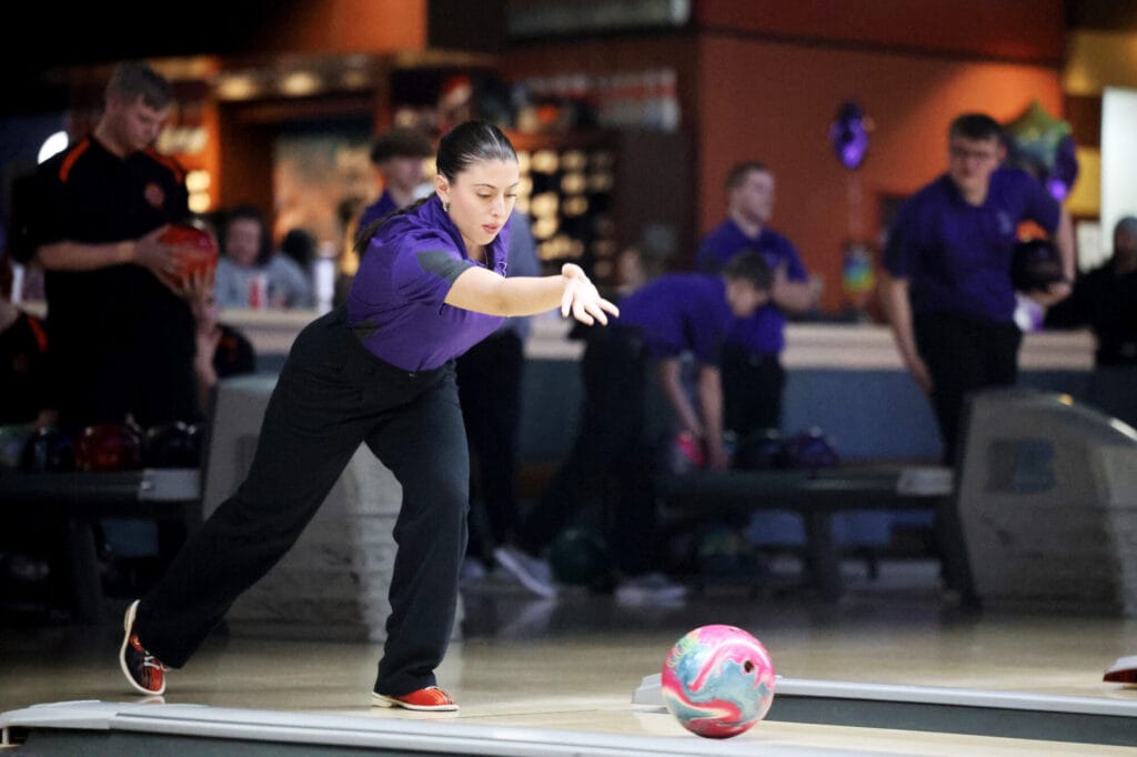 A member of the girls bowling team watches her ball roll down the alley after releasing it, her hand still in the air.
