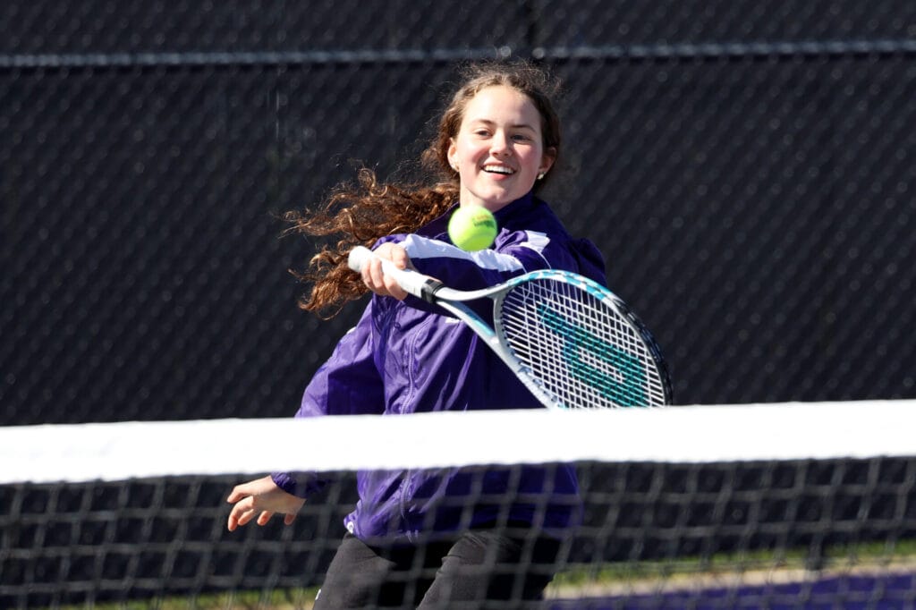 A member of the girls tennis team smiles while using her racket to hit a ball.