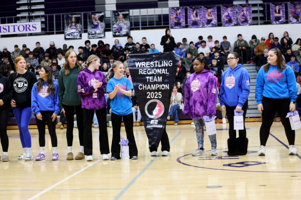 Members of the girls wrestling team are shown in the school gym while one of them holds up a banner reading "WRESTLING REGIONAL TEAM CHAMPION 2025."