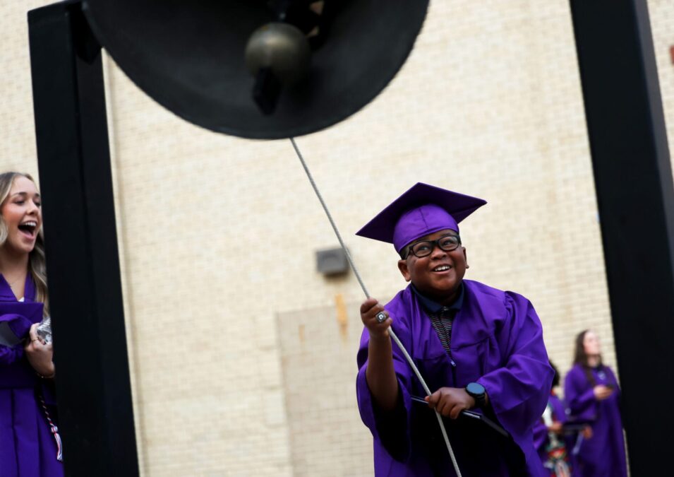 A young man wearing a cap and gown smiles while ringing the victory bell at Bracewell stadium following the commencement ceremony for the Burlington High School class of 2025.